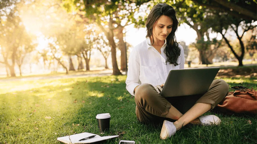A girl is sitting over green grass and working with a laptop, coffee, notebook & pen