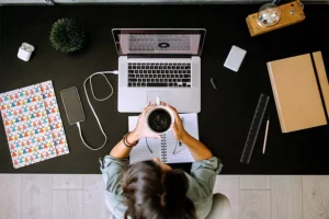 Top view of a person sitting at a desk holding a coffee cup in front of an open laptop, surrounded by notebooks, a smartphone, and other office supplies.