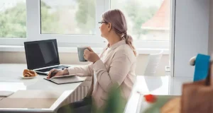 Woman with coffee looking out a window while working from home on a laptop, relaxed workspace with natural lighting