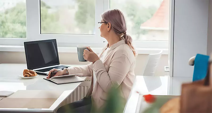 Woman with coffee looking out a window while working from home on a laptop, relaxed workspace with natural lighting