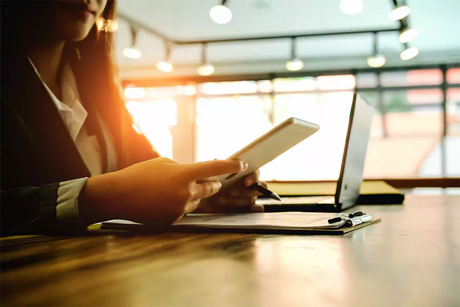 Person using a tablet and laptop while working at a desk, with a clipboard and sunlight in the background.