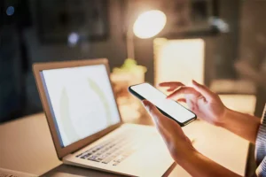 Person using smartphone while sitting at desk with laptop and desk lamp, symbolizing multitasking and digital work.