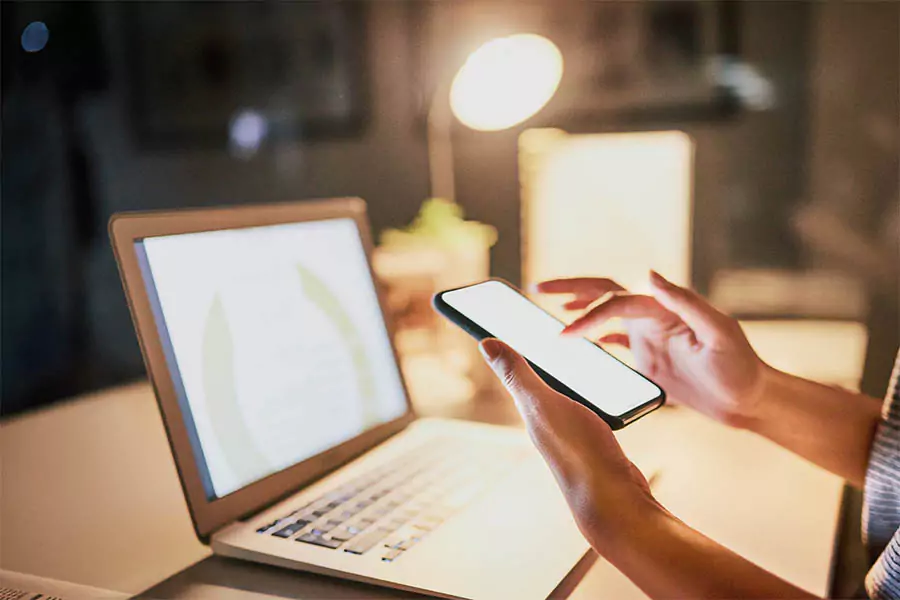 Person using smartphone while sitting at desk with laptop and desk lamp, symbolizing multitasking and digital work.